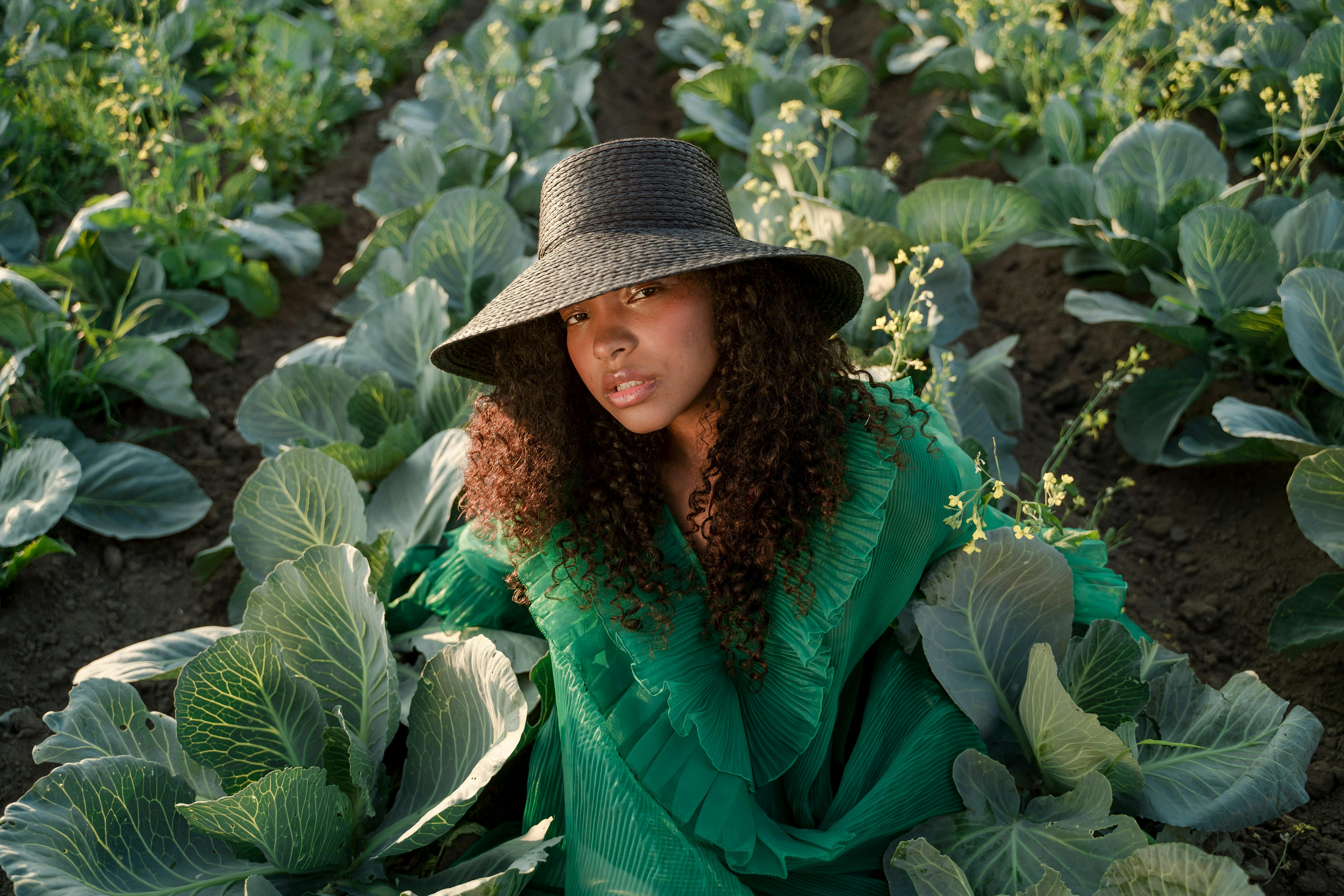 Woman Wearing Green Dress and Brown Hat Sitting Among Rows of Cabbages in Field