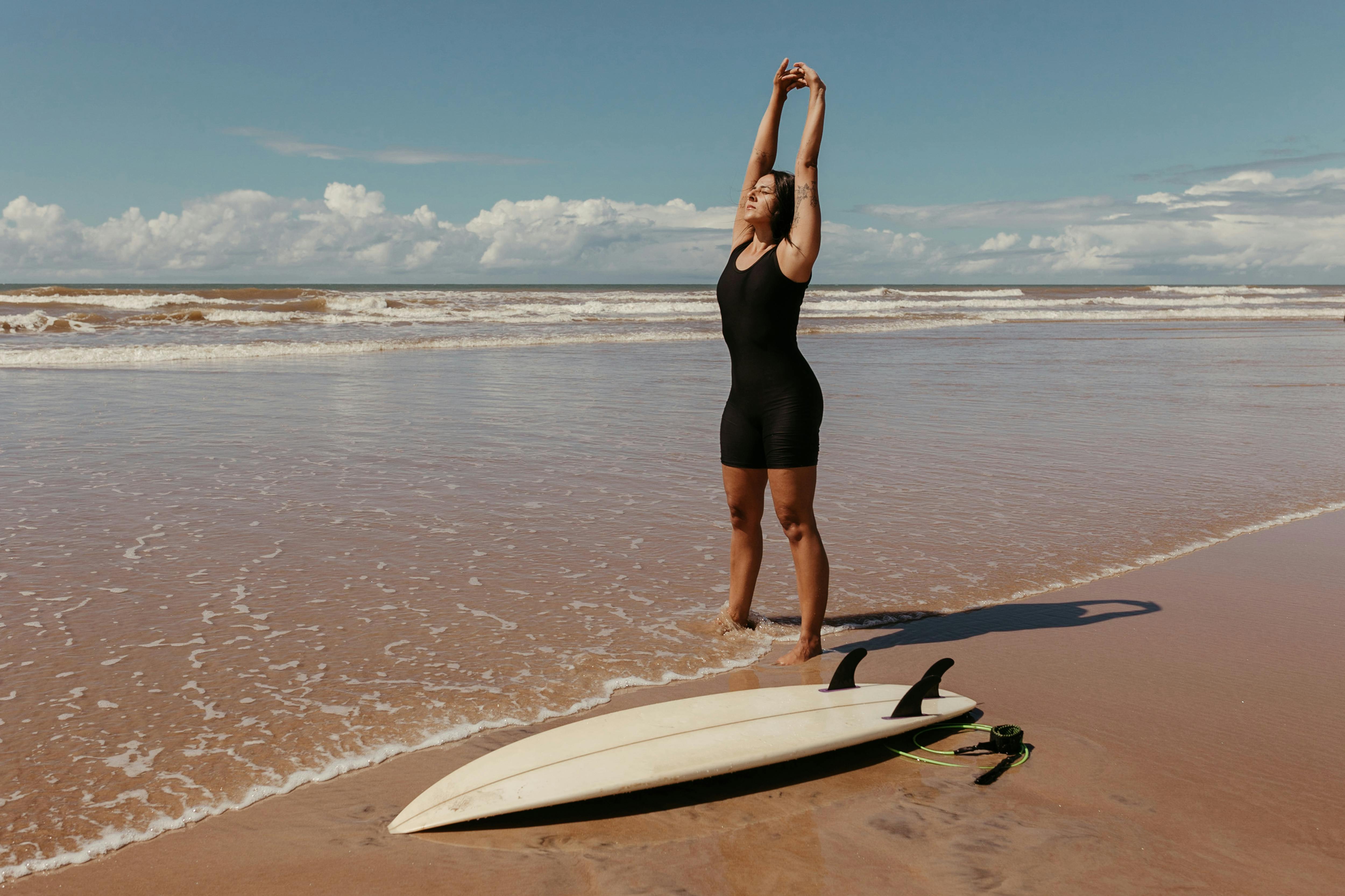 Surfer Stretching Next to Her Surfboard Standing in the Water on the Beach