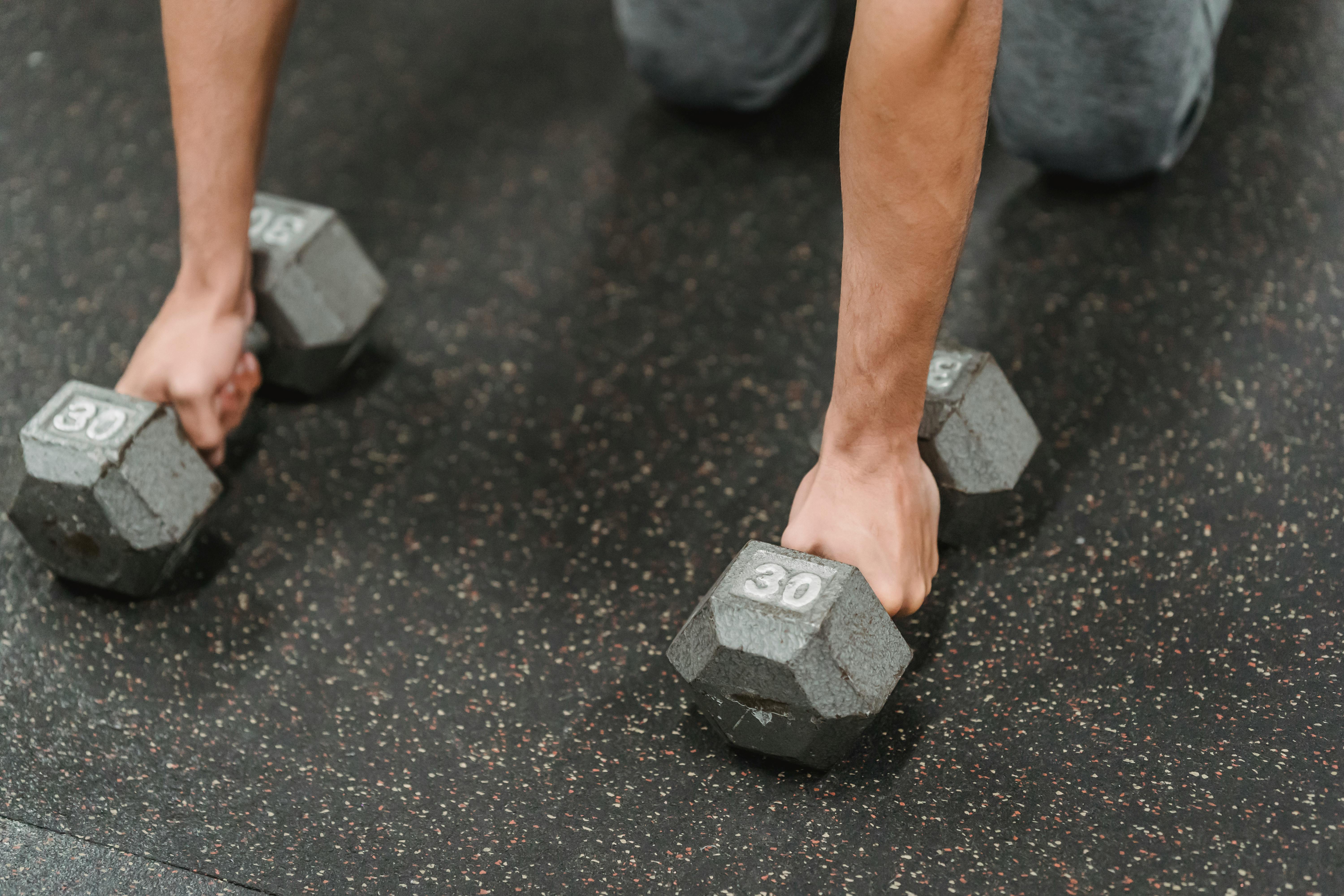 High angle of crop anonymous male athlete standing on knees with dumbbells while training on floor in fitness center