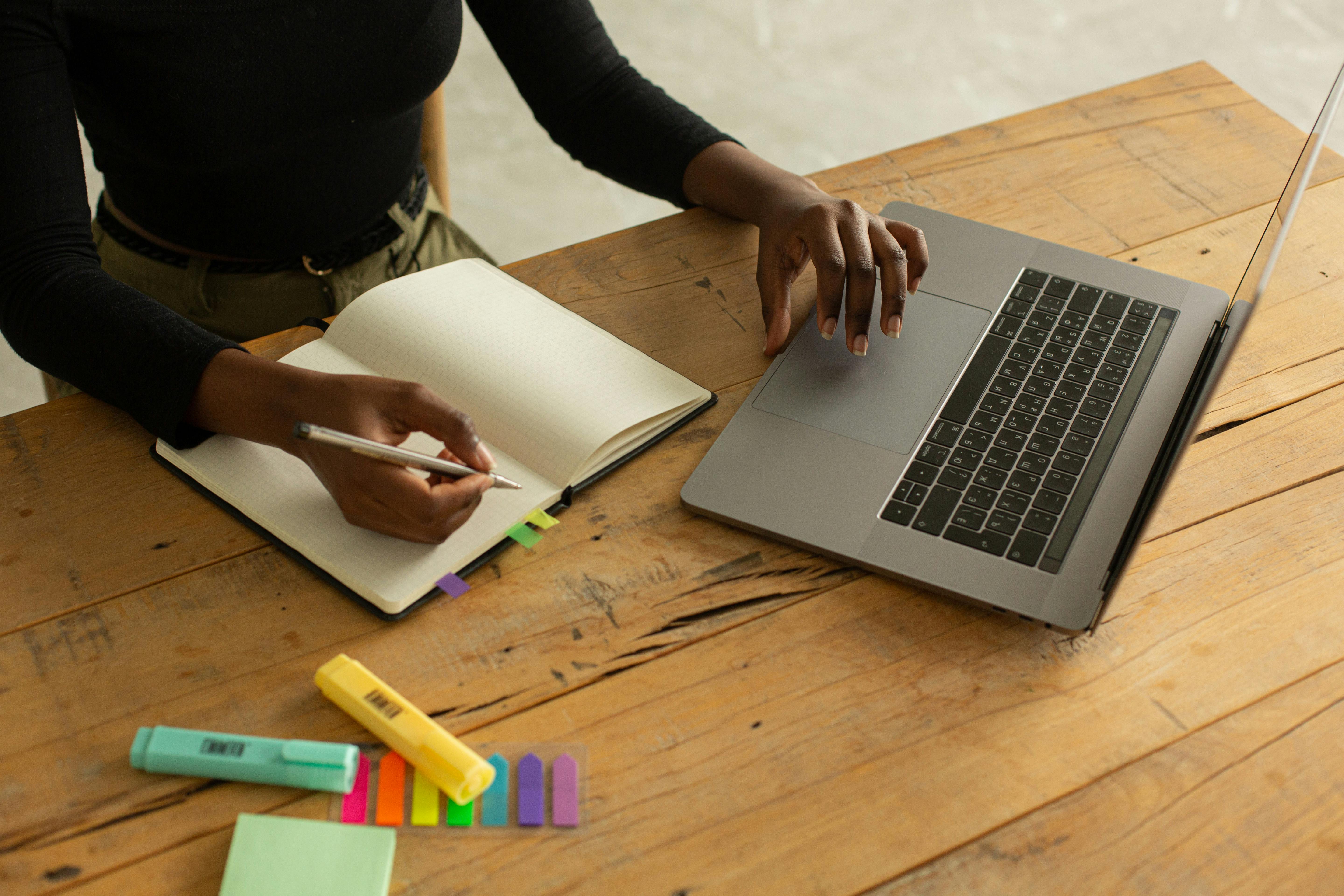 From above of crop black anonymous female freelancer sitting at table with opened laptop and notebook with markers and bookmarks and working remotely