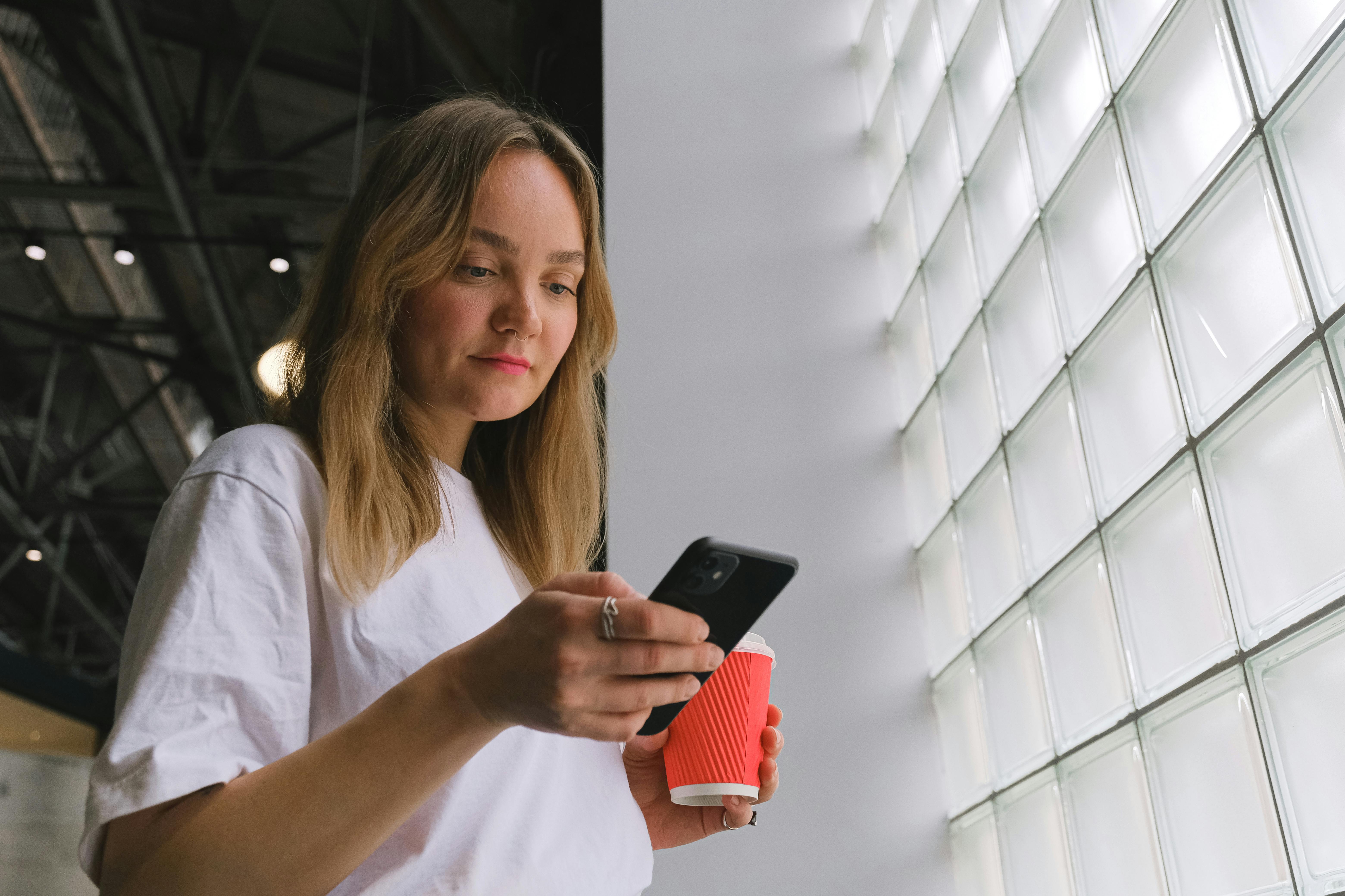 A Woman in White Shirt Using Her Phone while Holding a Cup of Coffee