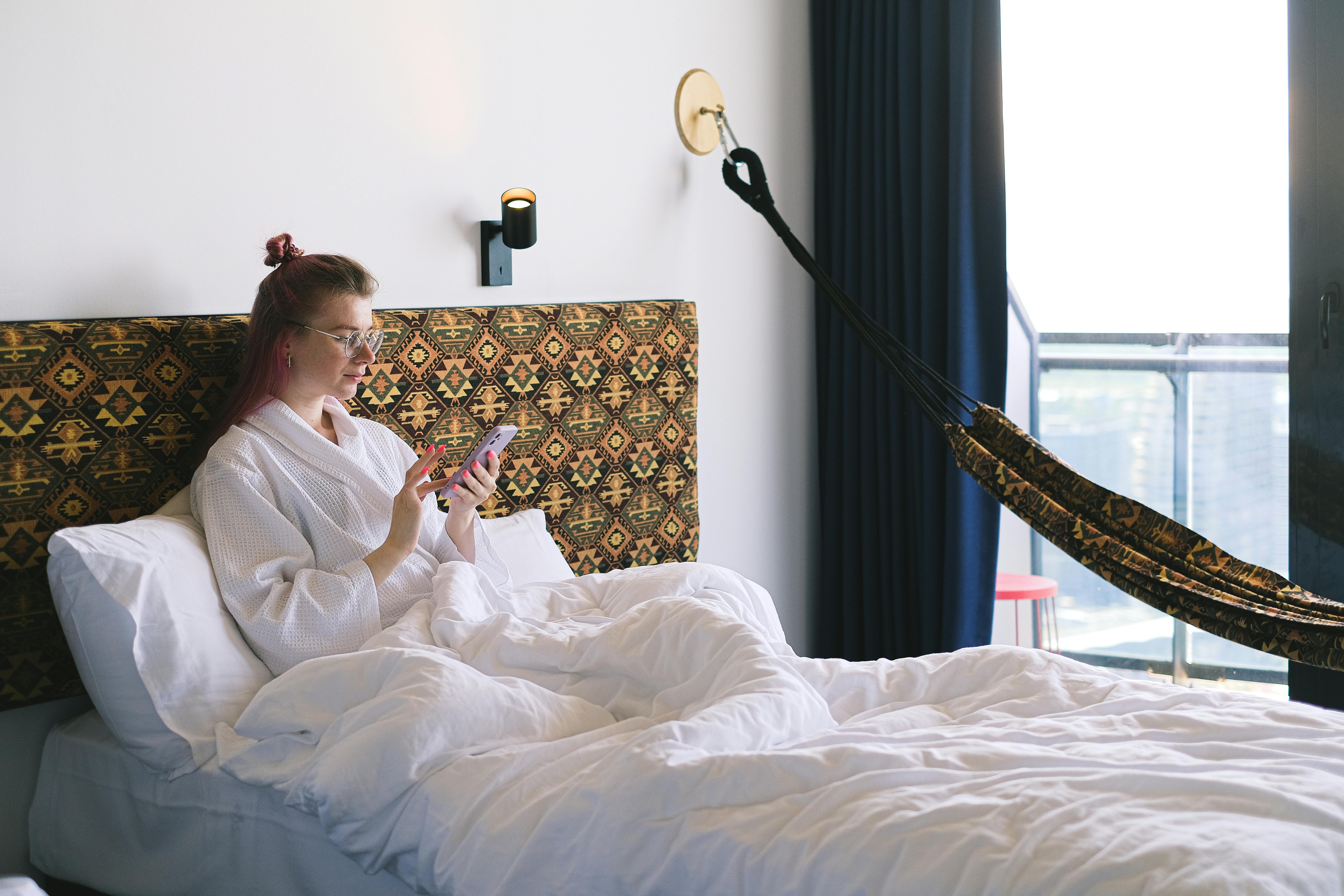 A Woman in White Bathrobe Sitting on the Bed while Using Her Phone