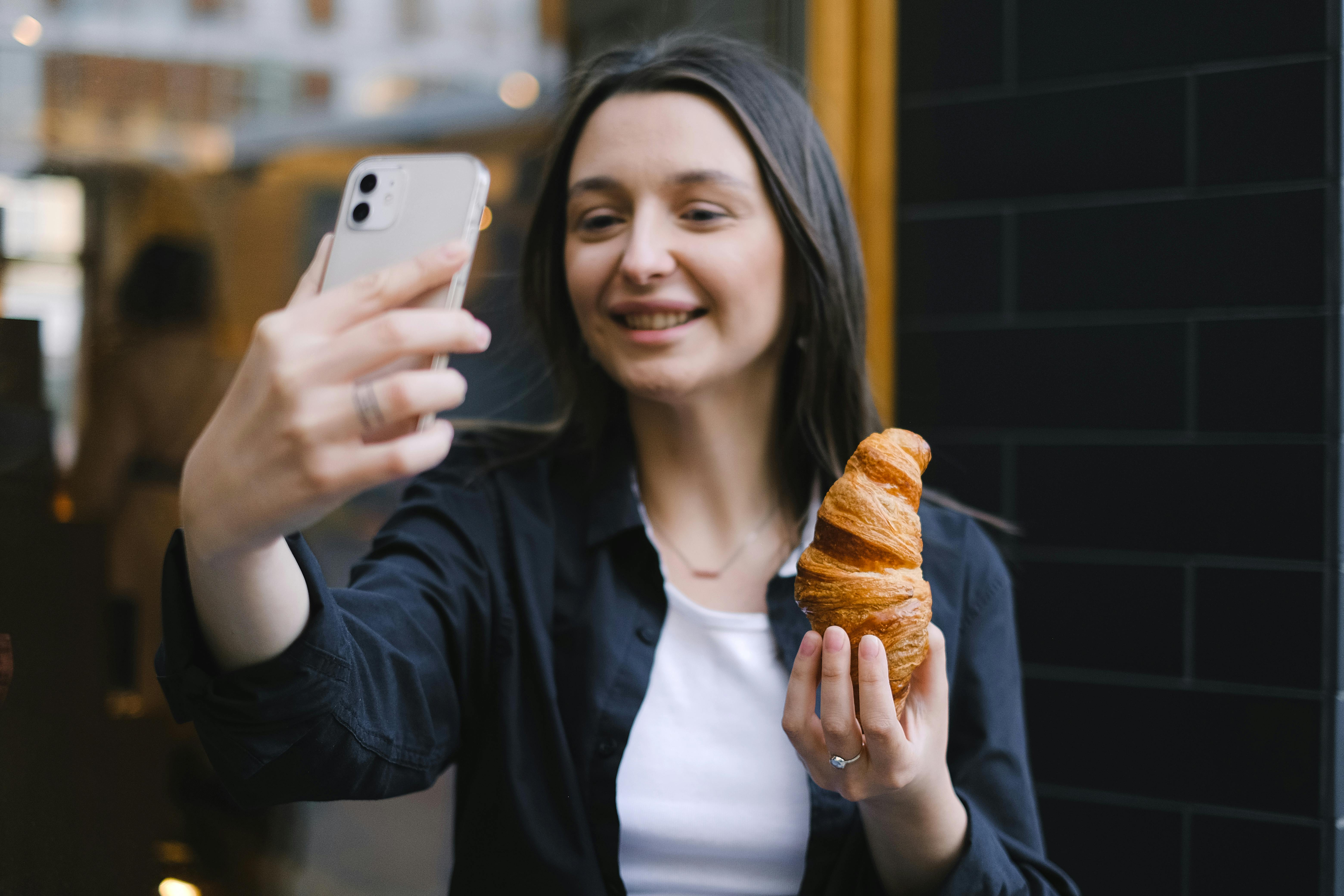 Smiling Woman Holding a Croissant and a Smartphone