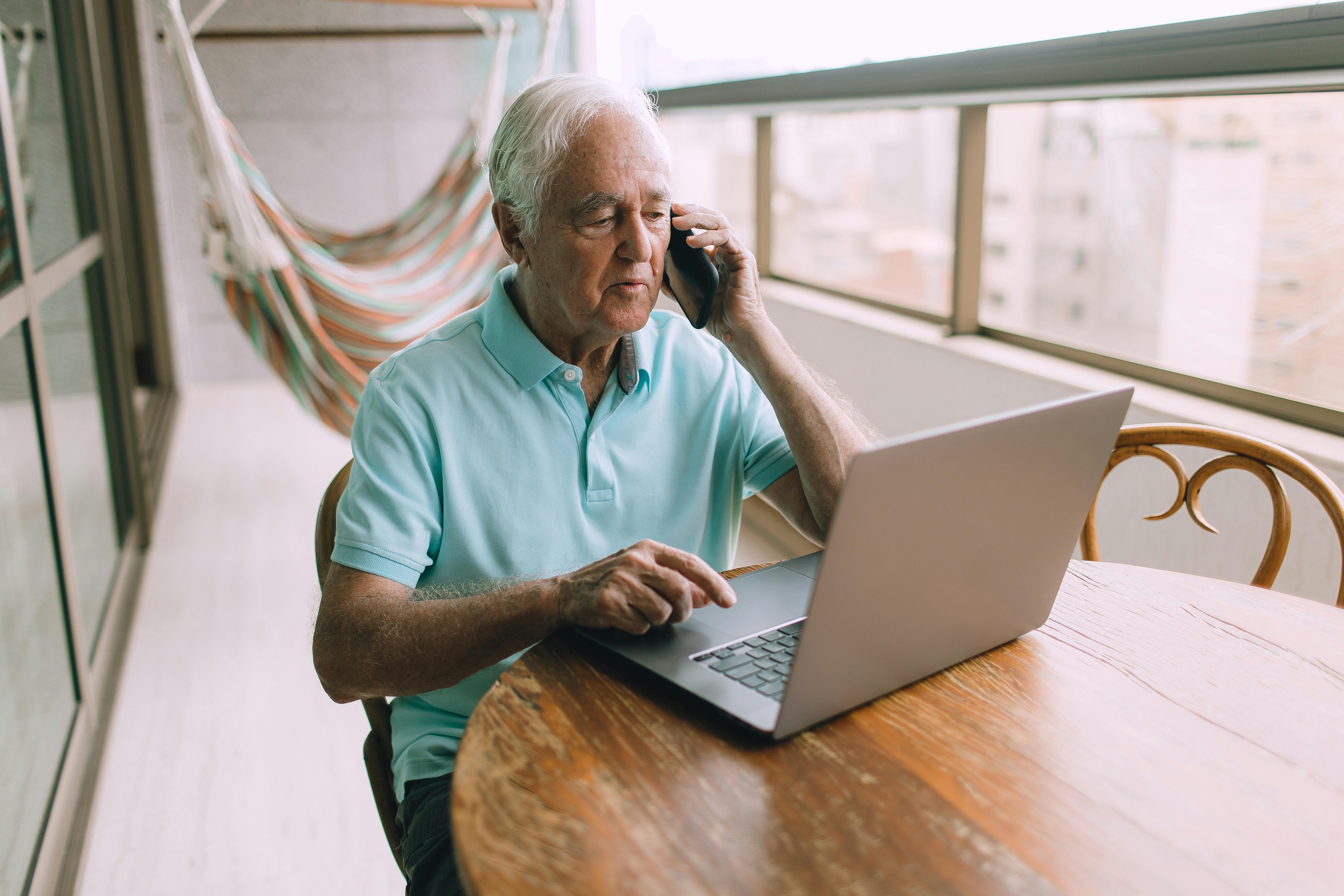 An Elderly Man Talking on the Smartphone by the Laptop