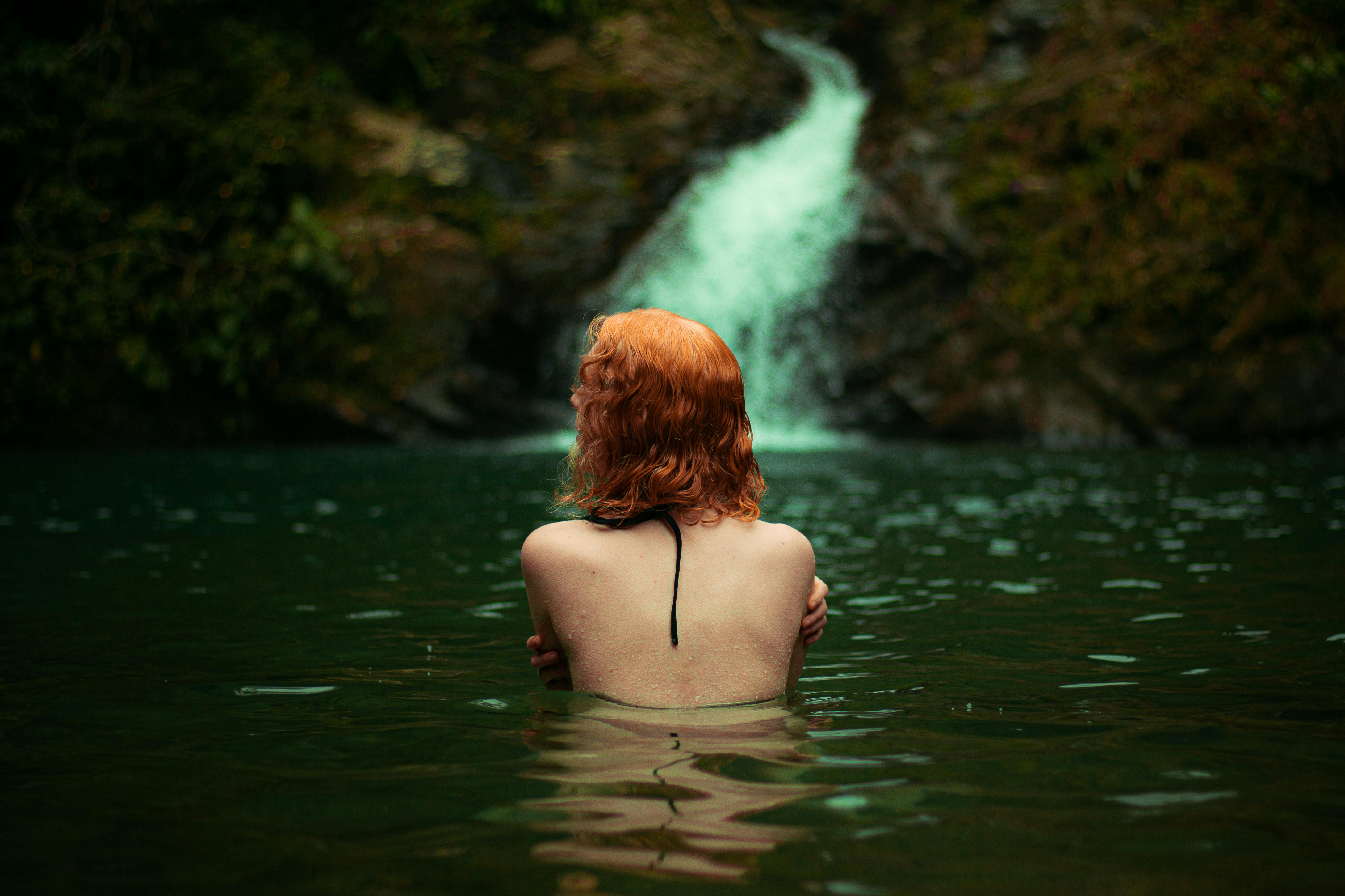 A woman with red hair in the water near a waterfall