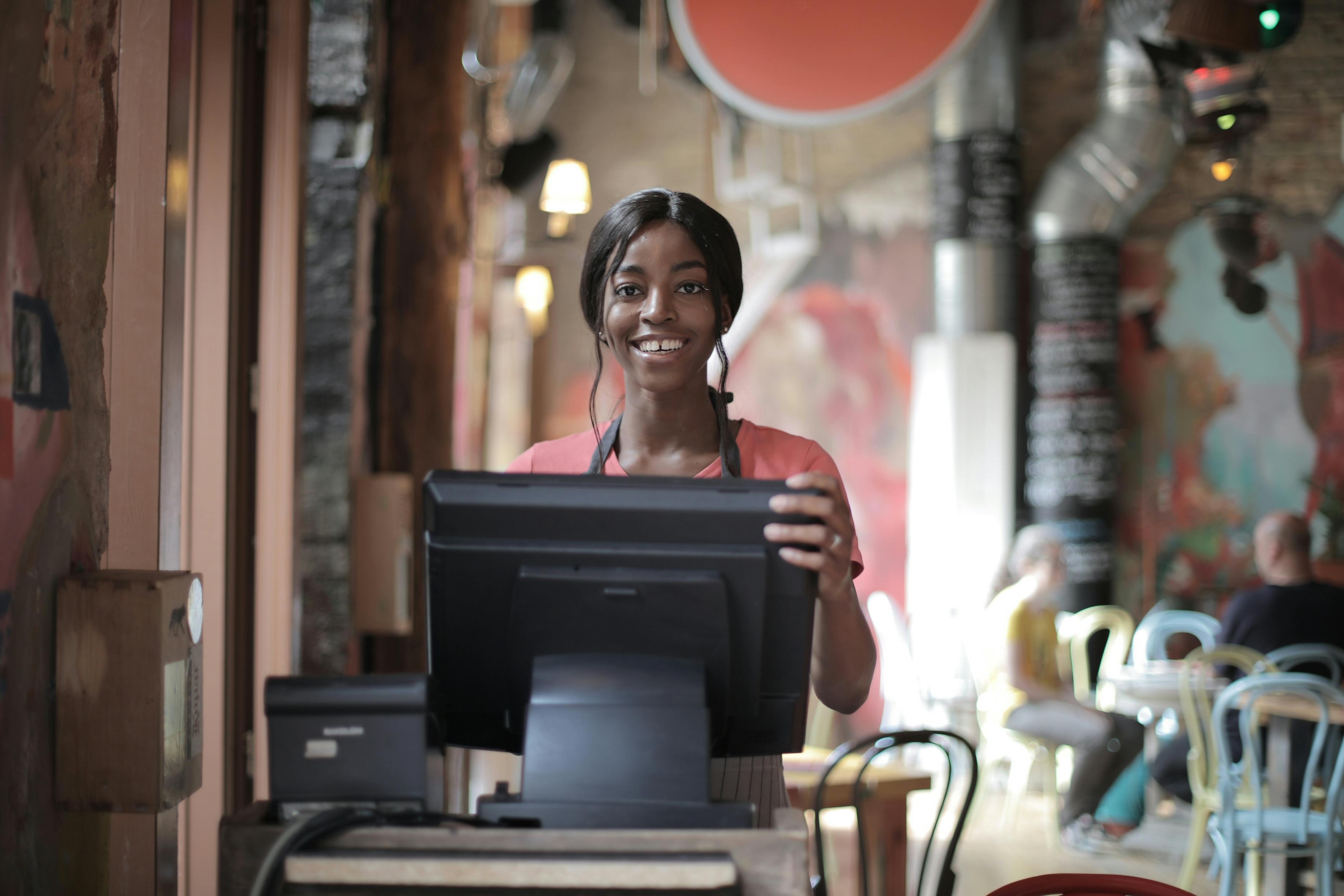 Positive young woman in uniform smiling while standing at counter desk in  cafe