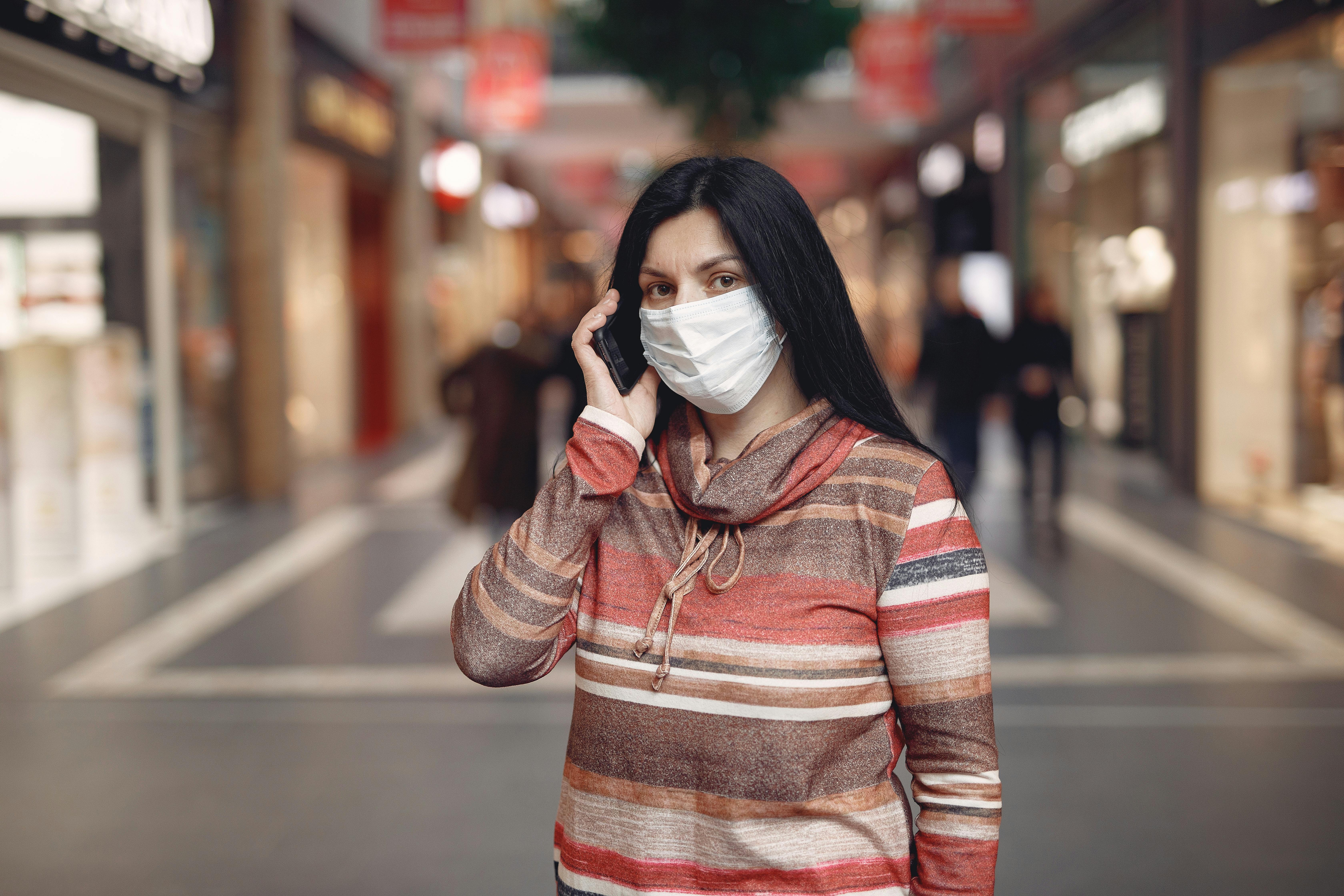 Worried casual female wearing protective mask and looking at camera while answering phone call against blurred background in city center during coronavirus pandemic