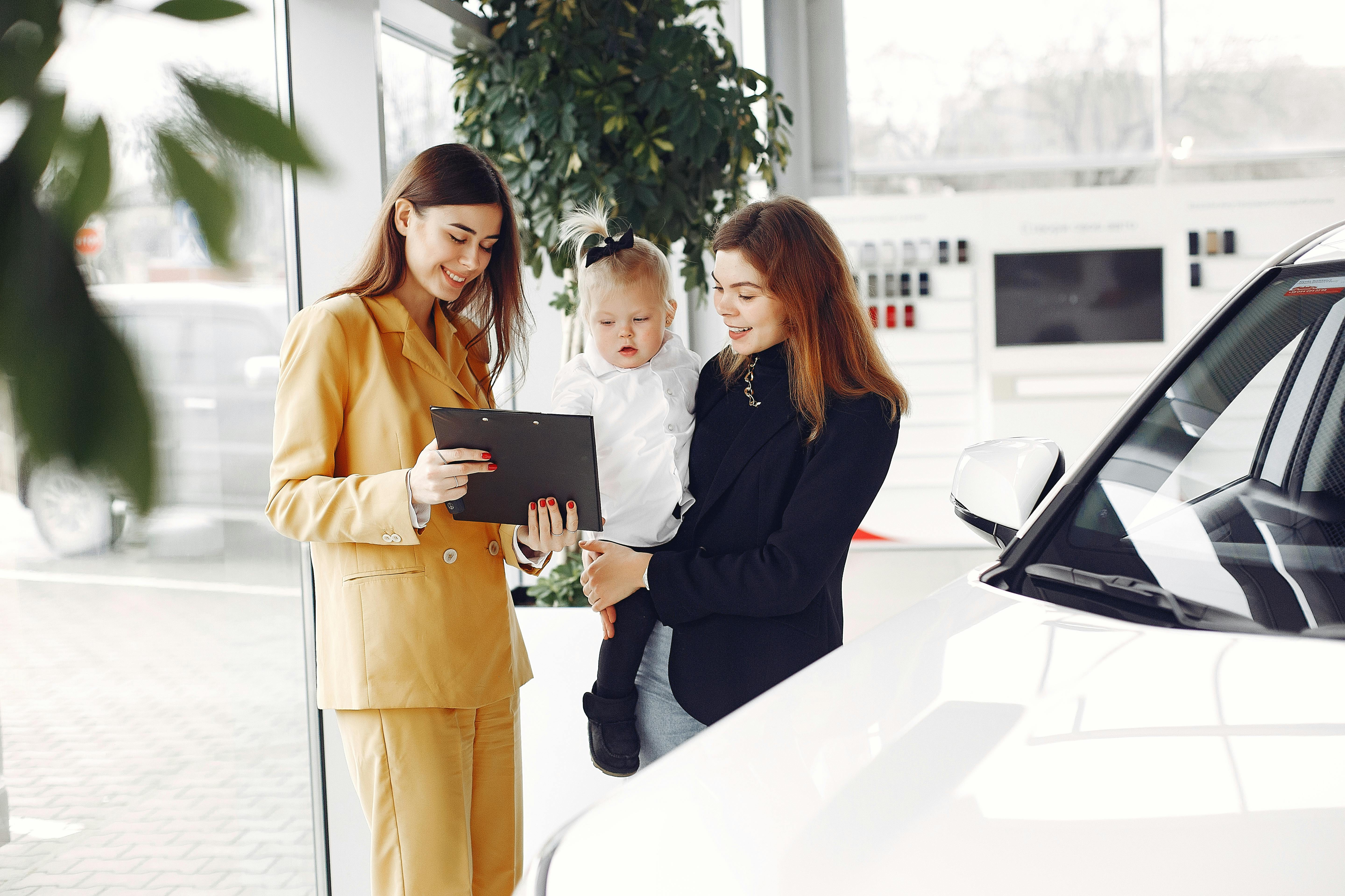 Positive young female with cute little daughter discussing car characteristics with professional dealer in stylish beige suit while standing in car showroom in daylight