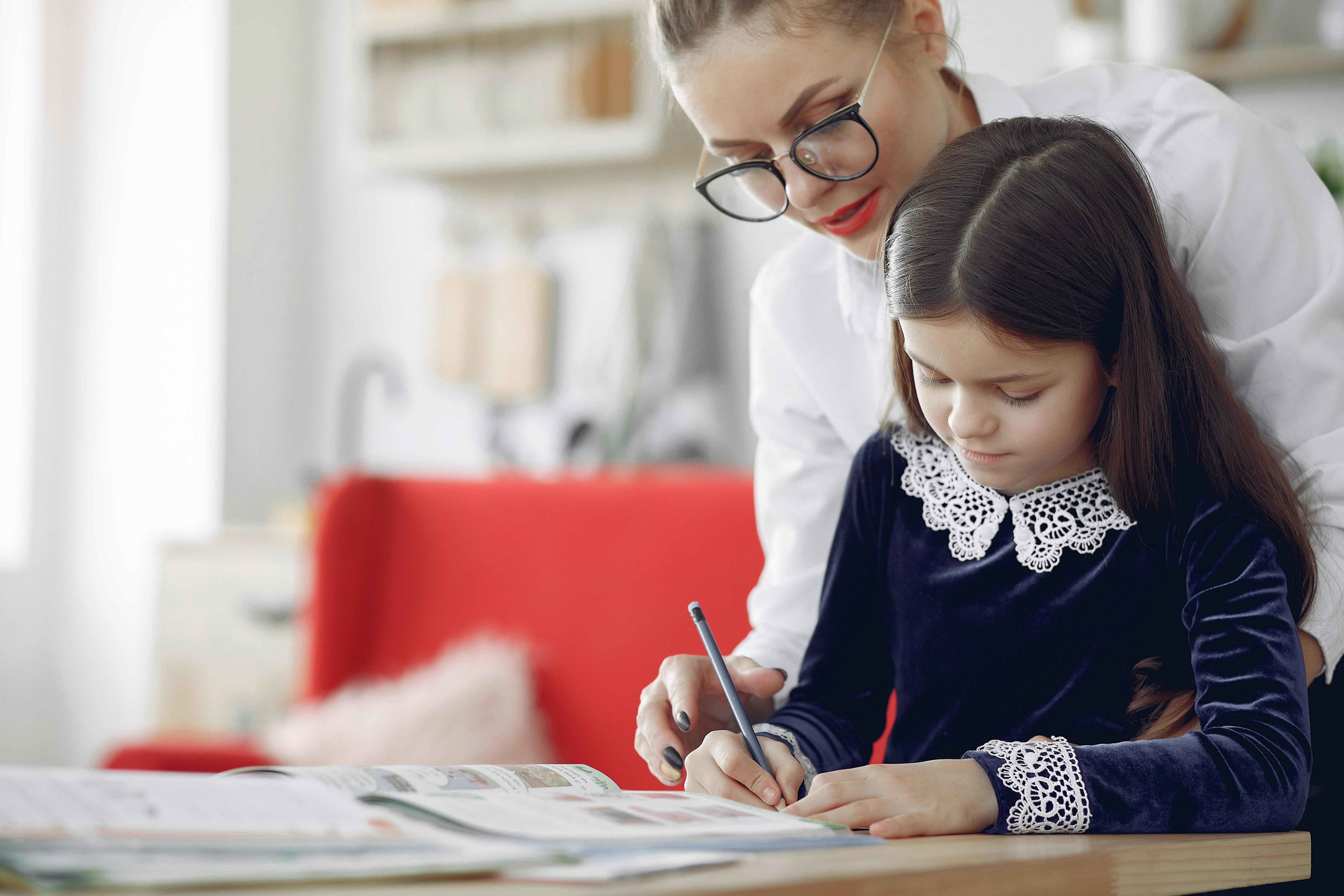 Young girl in casual clothes sitting at table and writing with tutor while working on homework assignment together in contemporary apartment