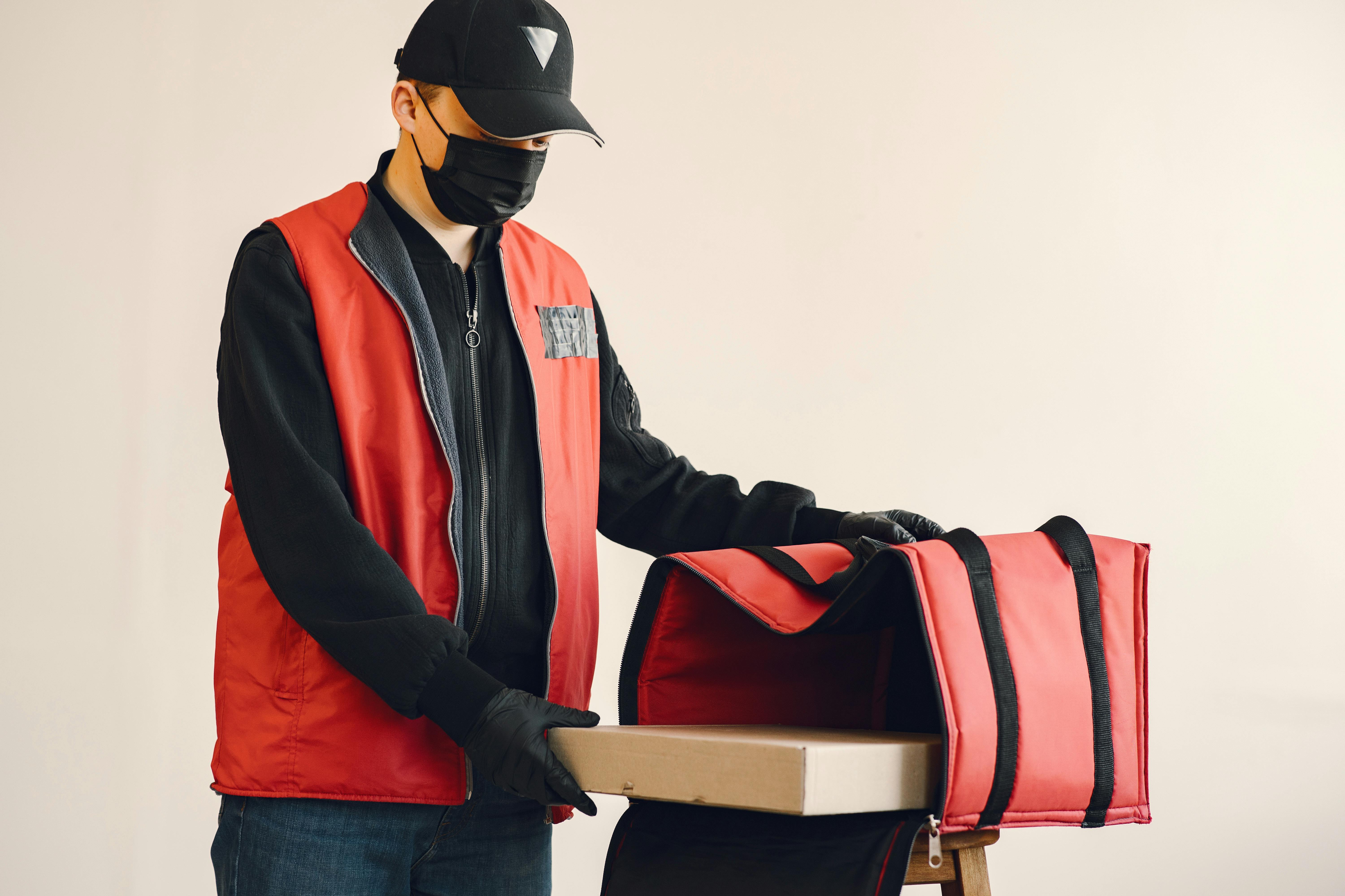 Young delivery man wearing surgical facial mask and gloves for preventing disease during coronavirus pandemic putting pizza box in thermo bag for safety food delivery