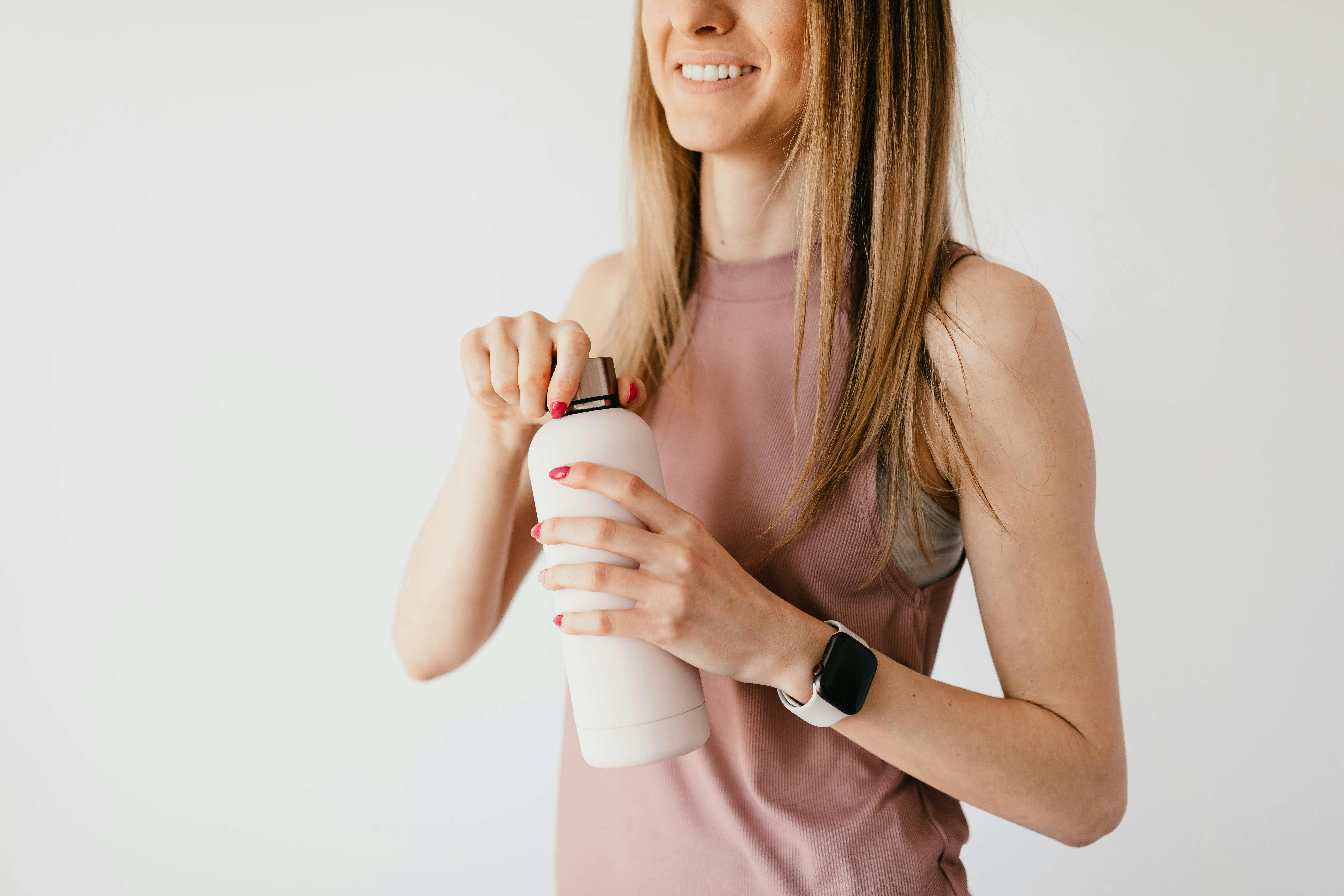 Crop faceless young female in casual outfit wearing smart watch opening cosmetic bottle while standing against white background