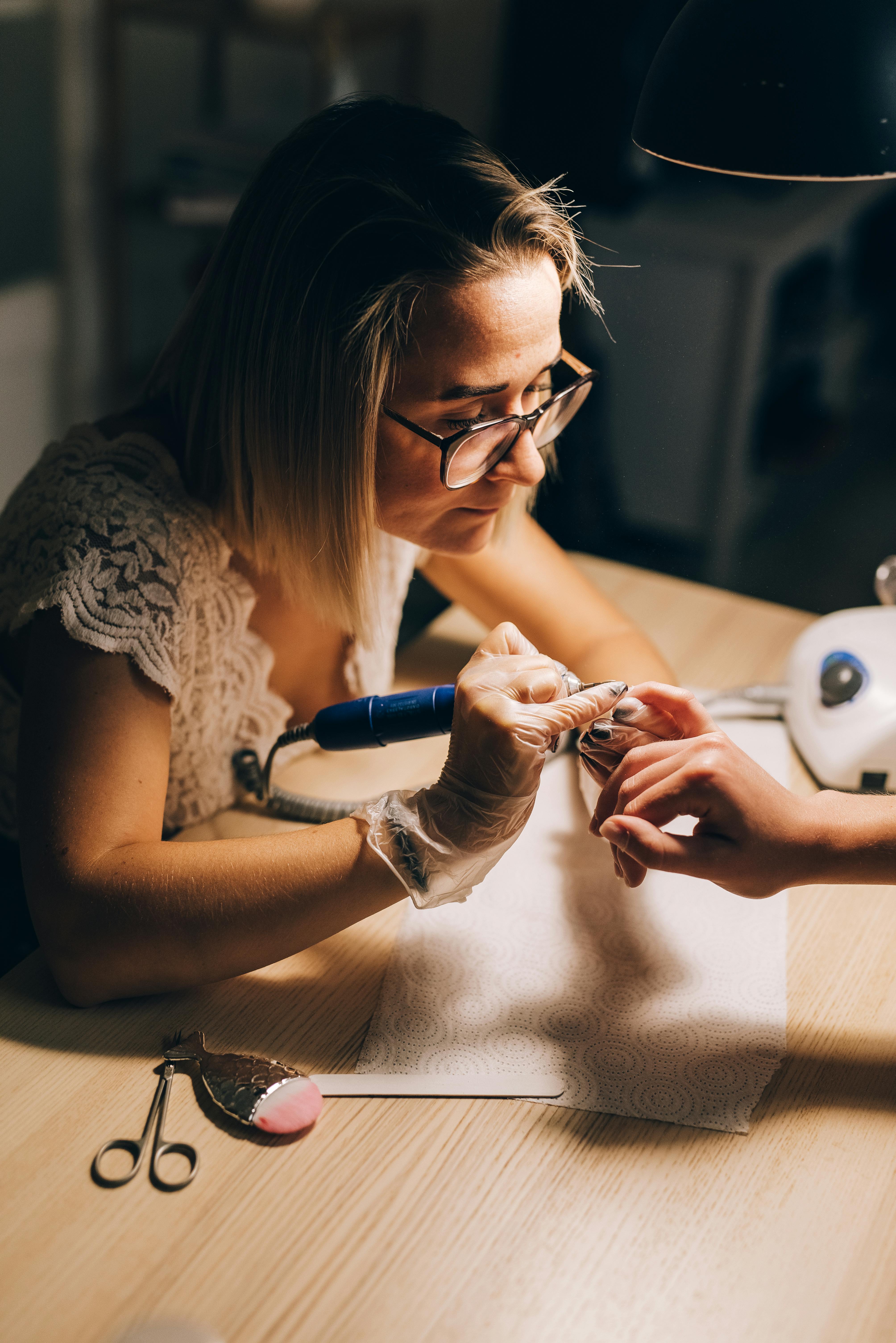 High angle of concentrated female master in glasses and gloves manicuring hand with drill of faceless client