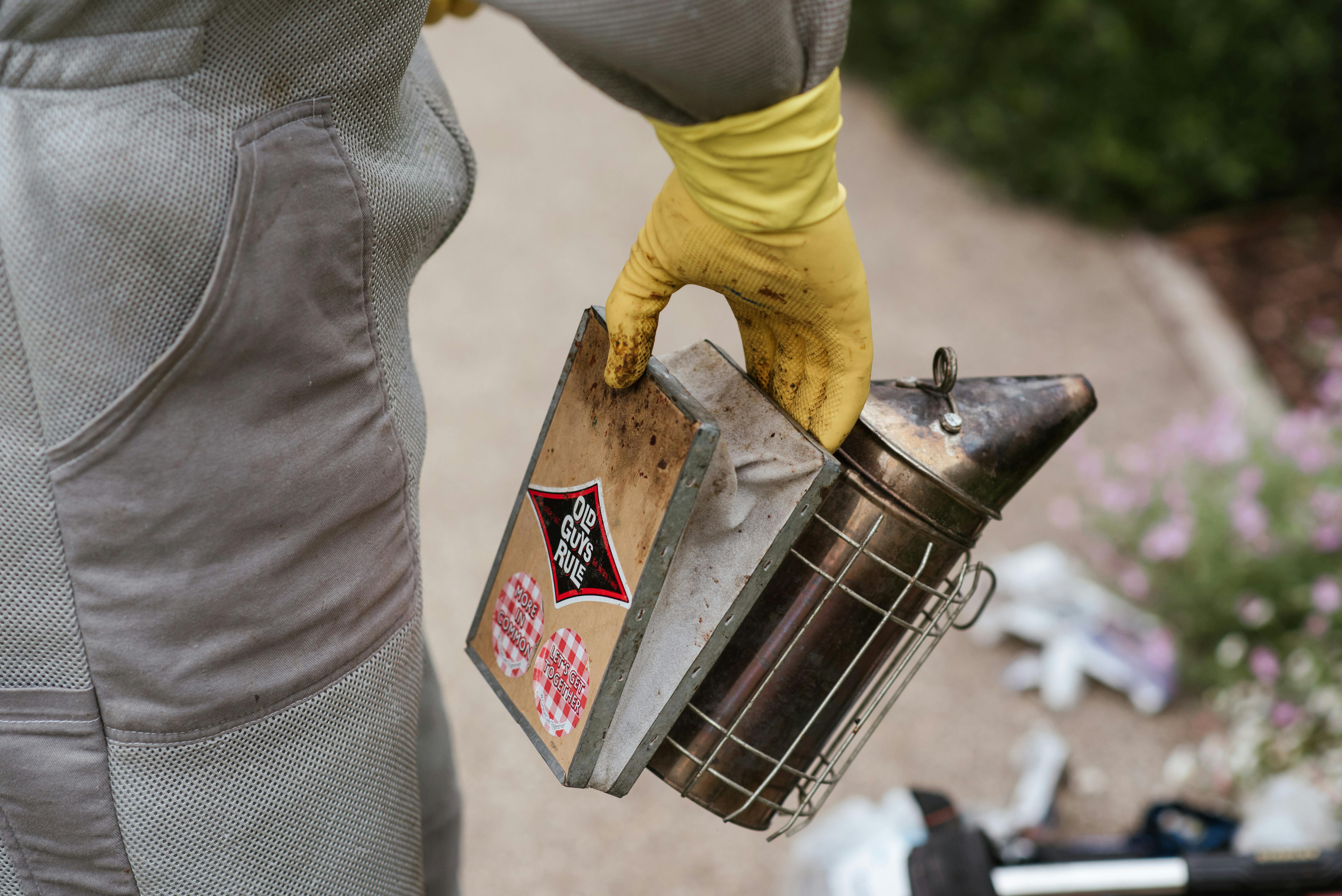 Crop faceless male beekeeper in gloves and protective uniform standing in countryside and holding bee smoker