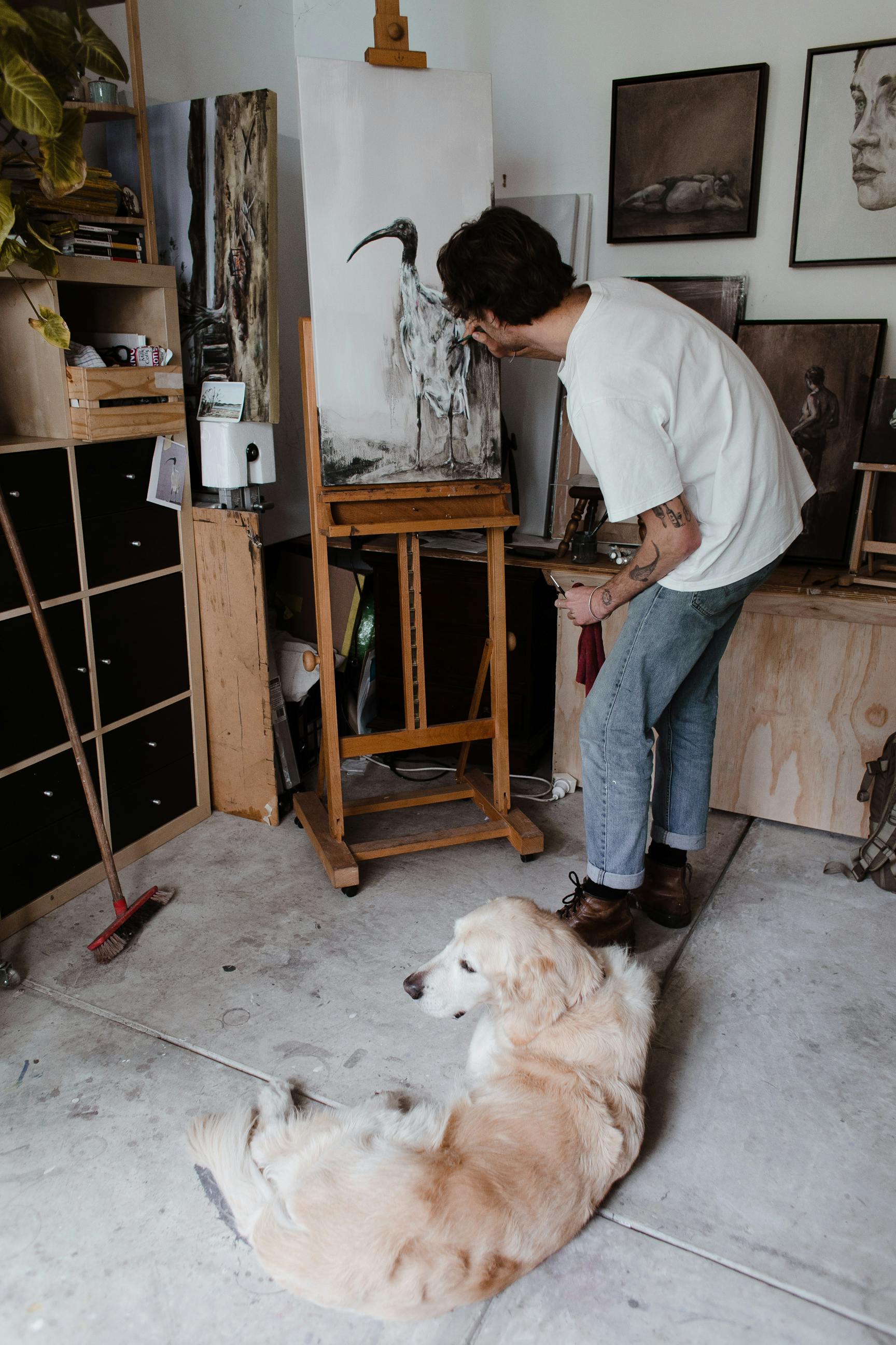 Side view of unrecognizable young male artist in casual clothes painting on easel while standing in light workshop near adorable Golden Retriever dog