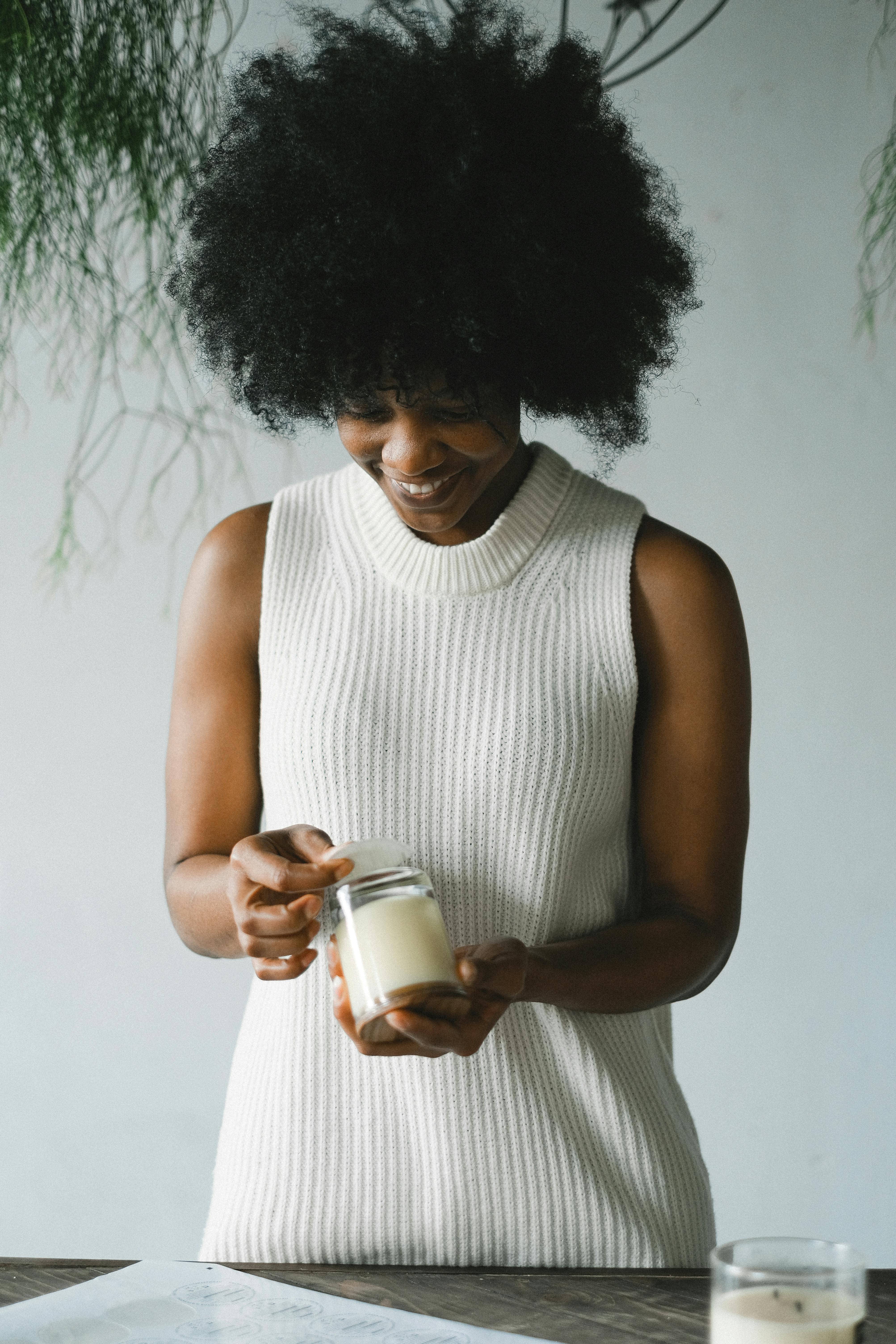 Positive African American female artisan standing at table with handmade candles in glasses in workshop