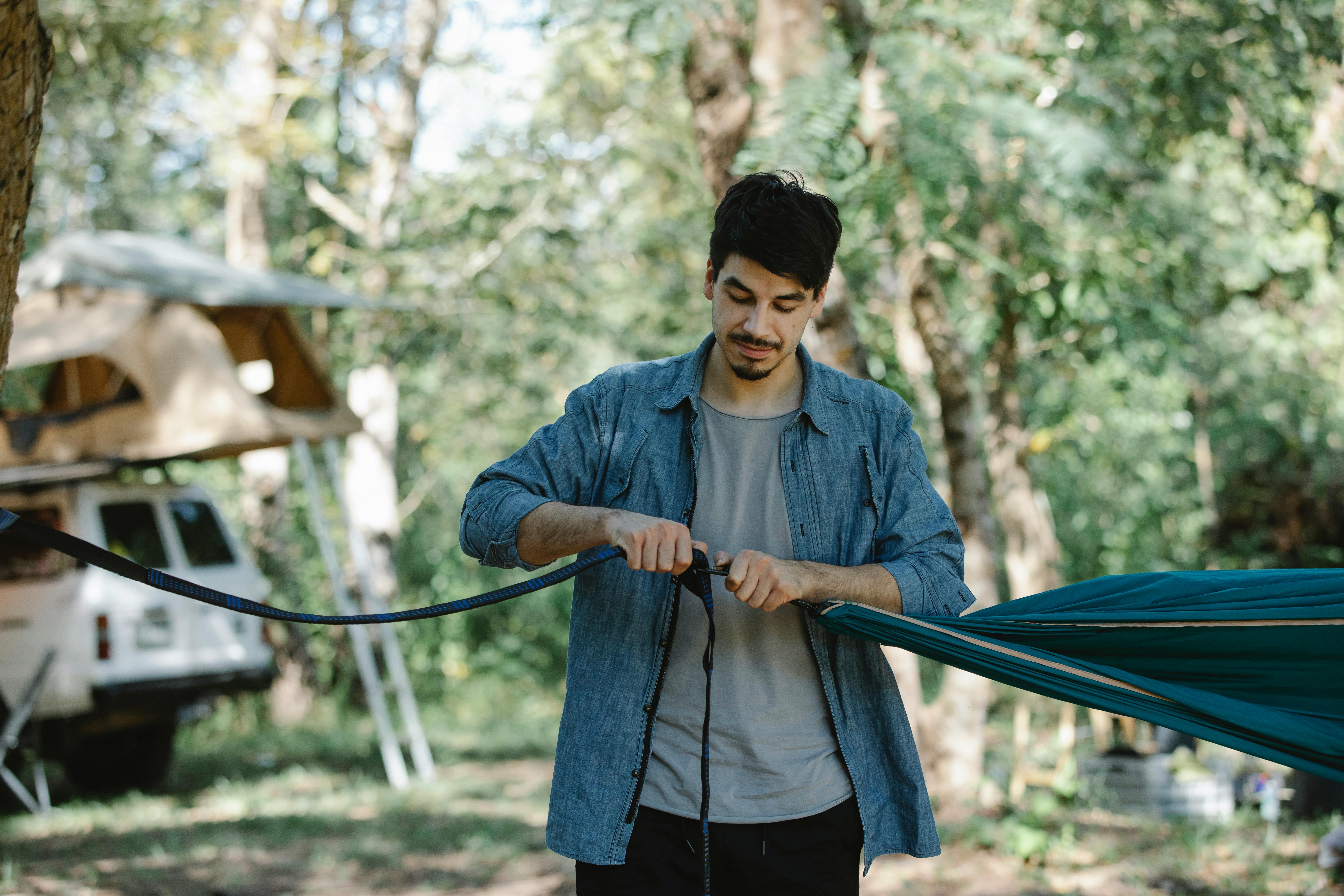 Focused young bearded male traveler hanging hammock between trees in forest while preparing camp against car and tent