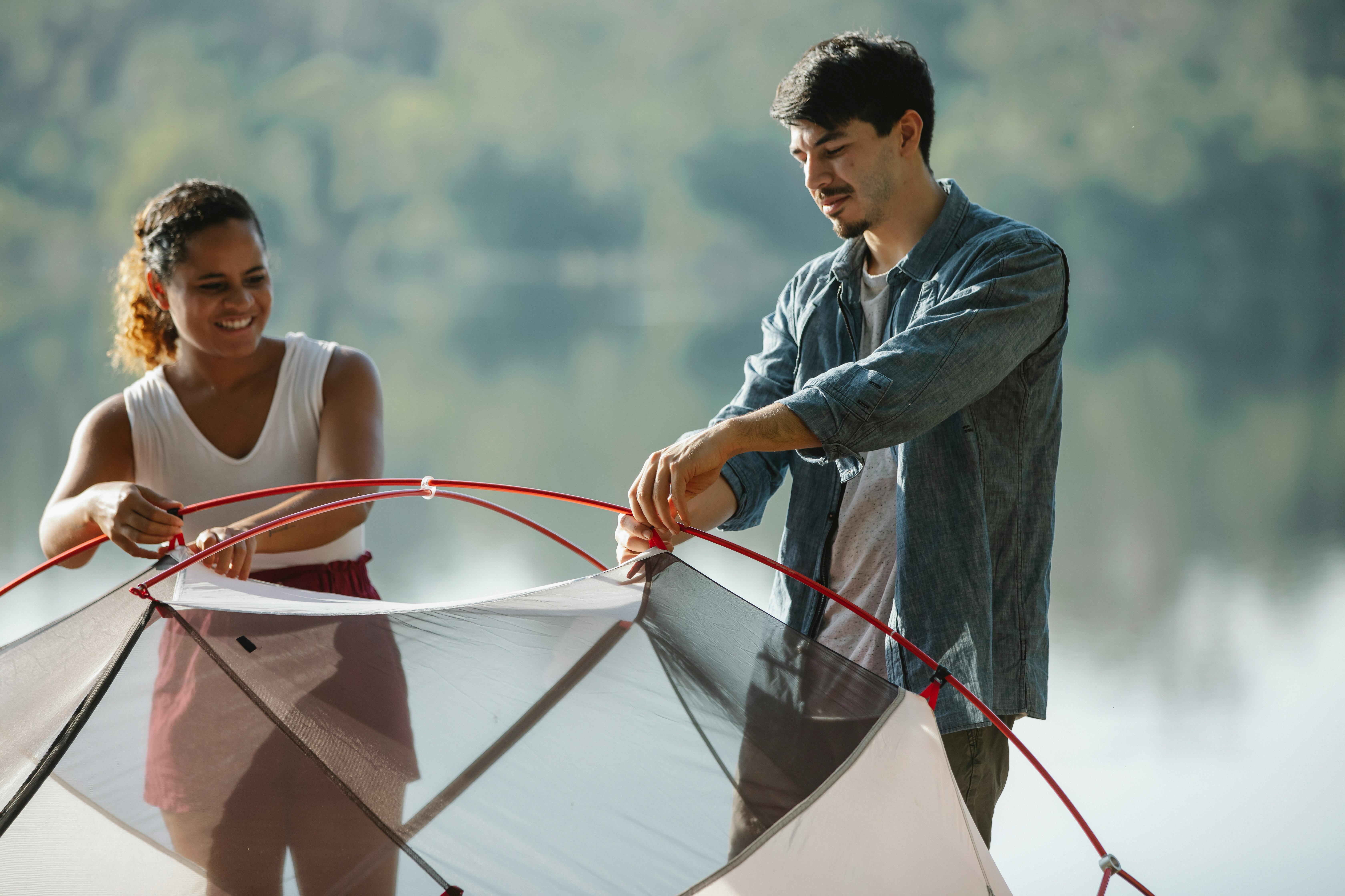 Young diverse traveling couple in casual outfits putting up tent near lake in summer day