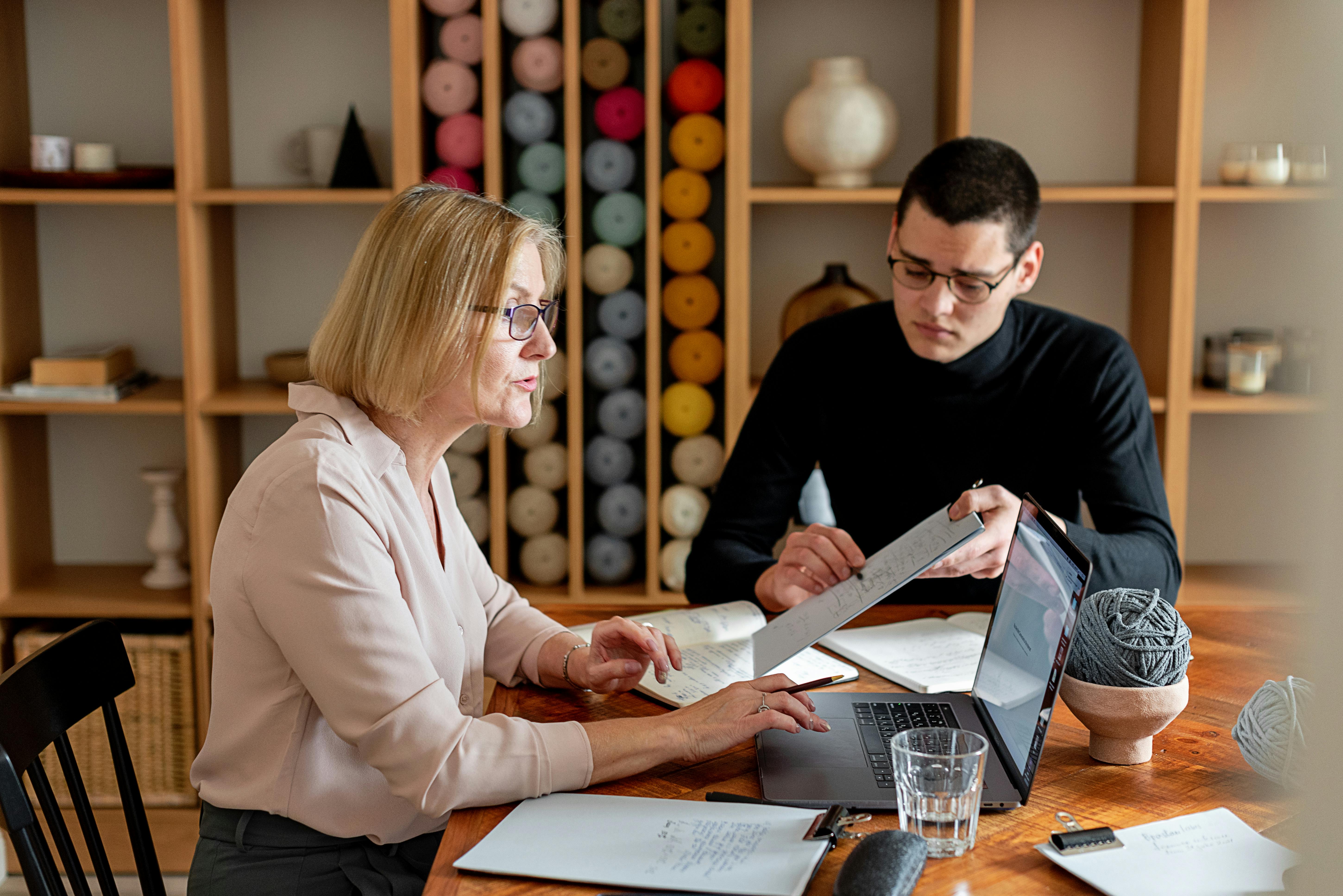 Man in Black Long Sleeves Showing Papers to Woman in Beige Dress Shirt Using Laptop