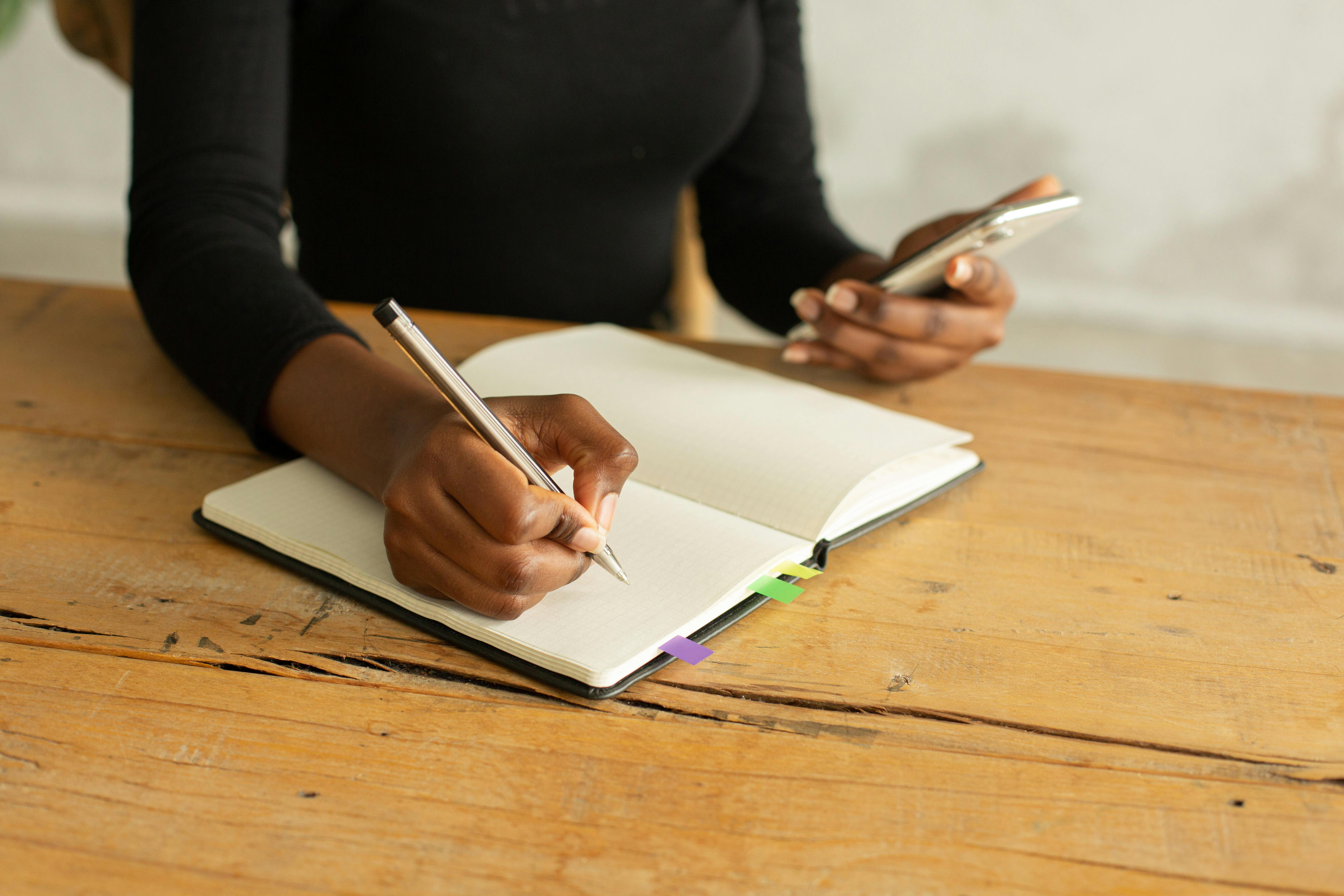 Crop anonymous ethnic female sitting at table and taking notes on white blank pages of notepad while looking at screen of mobile phone