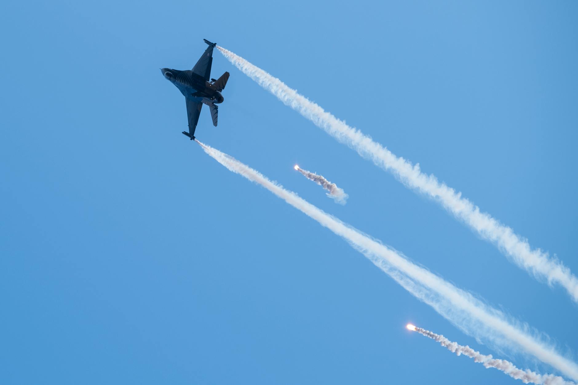 A fighter jet performing complex maneuvers with flare trails in a clear blue sky.