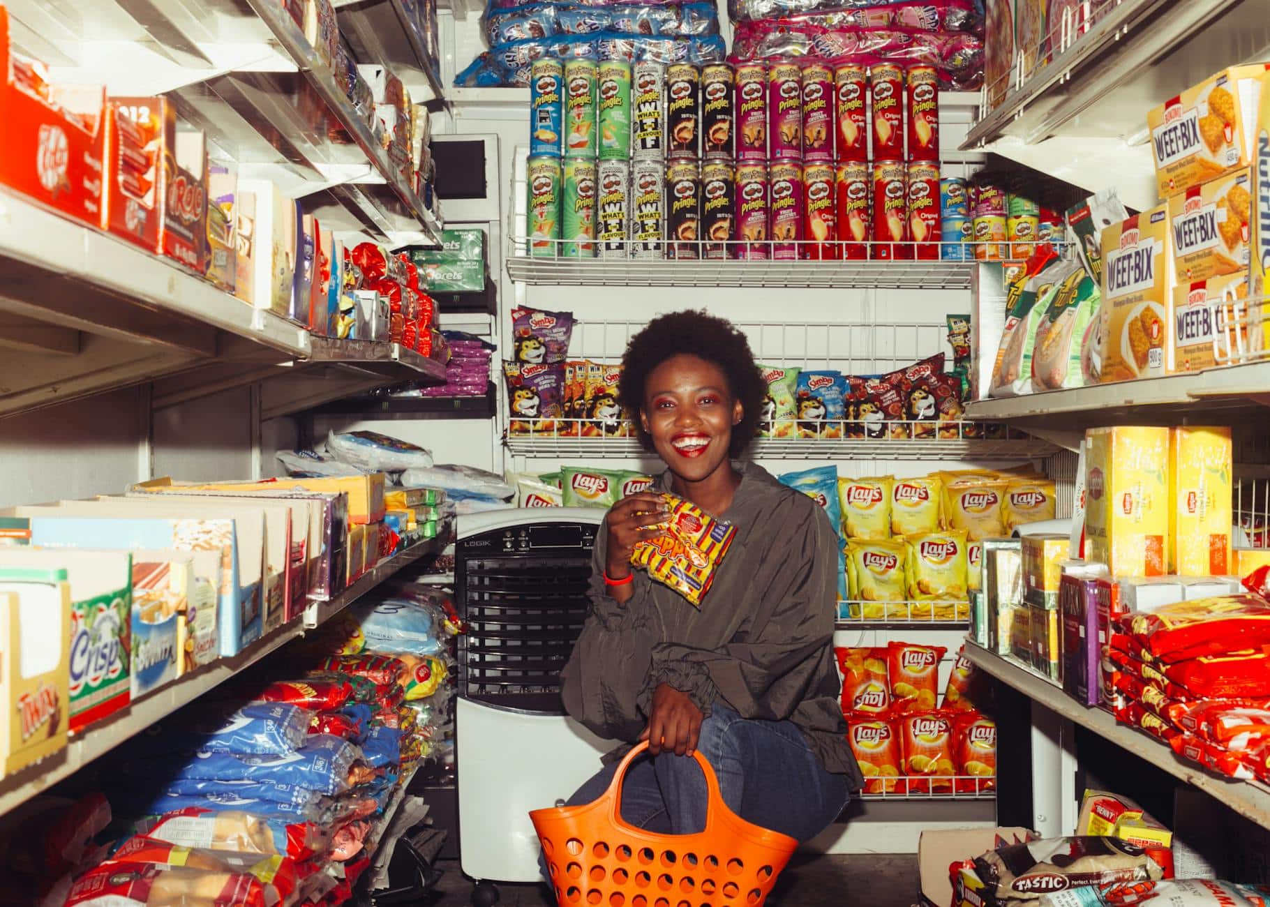 Happy black female in casual clothes with shopping bag demonstrating product squatting among grocery shelves while choosing goods in supermarket