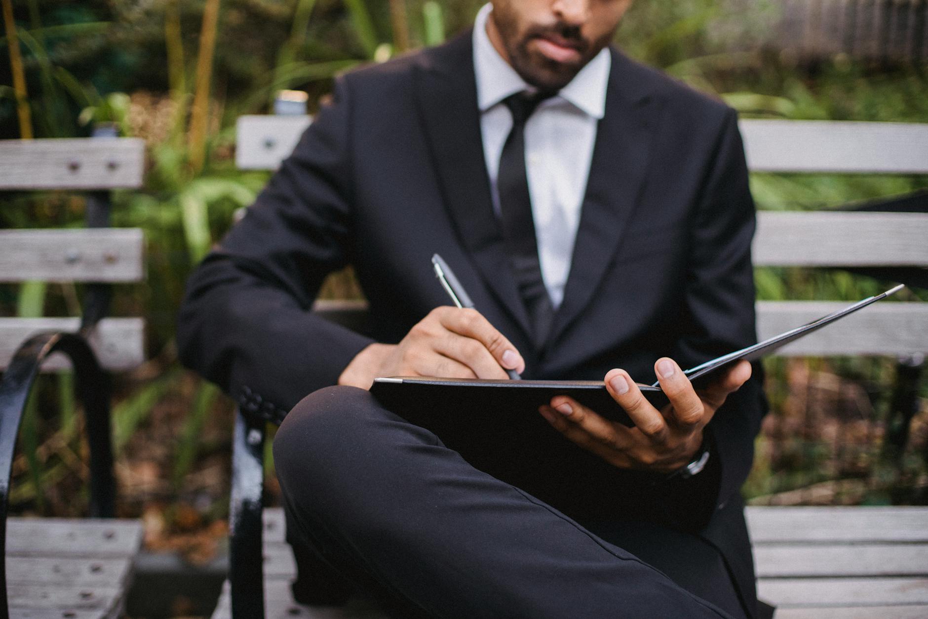 A Man in a Black Suit Writing While Sitting on a Bench