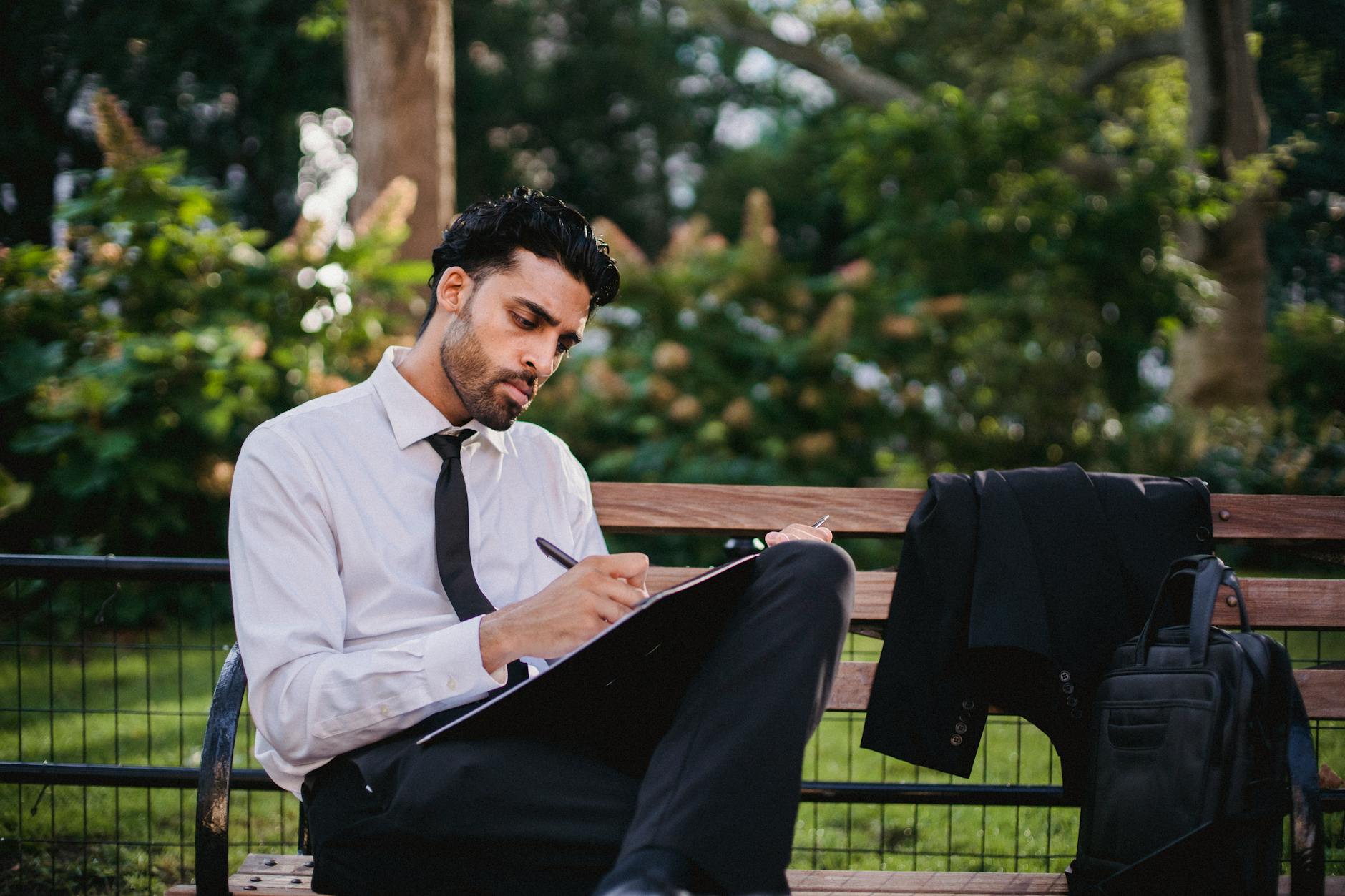 A Man in a White Long Sleeves and Black Pants Writing While Sitting on a Brown Wooden Bench