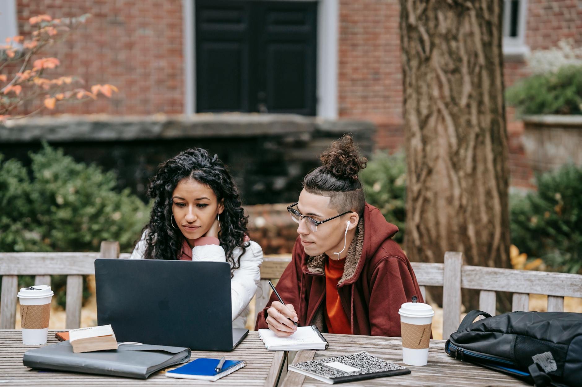 Young multiethnic friends studying while using netbook at table with coffee and planners in yard of brick building