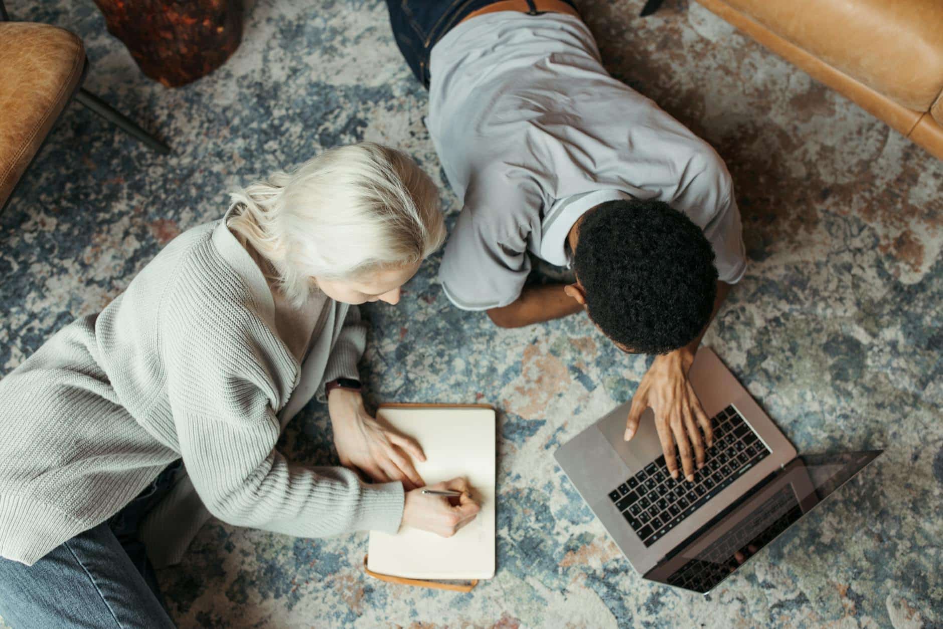 Man Using a Laptop Beside a Woman Reclining on Floor Carpet and Writing on a Paper