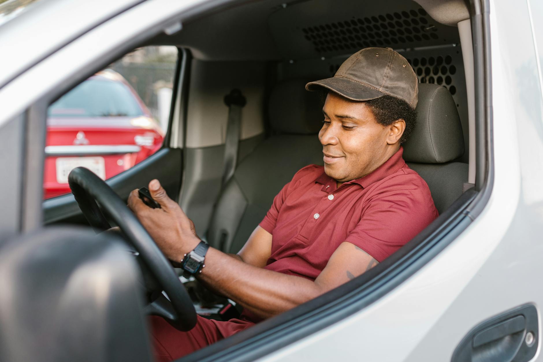 A man in a red polo shirt and brown hat driving a car.