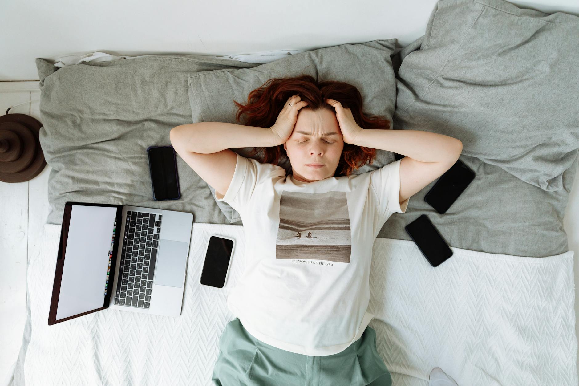 A Stressed Woman Lying on a Bed beside Cellphones and a Laptop